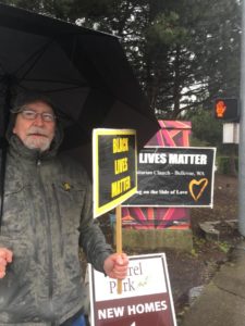 A man under an umbrella in a heavy coat stands proudly in front of and holding signs in support of black lives.